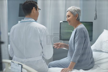 Foto de Vista trasera del joven médico de bata blanca discutiendo el estado de salud del paciente mayor mientras está sentado en la cama en la sala de hospital - Imagen libre de derechos