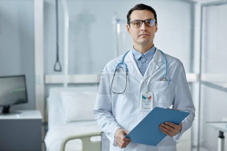 Foto de Retrato del joven médico de bata blanca con tarjeta médica mirando a la cámara mientras está de pie en el hospital - Imagen libre de derechos