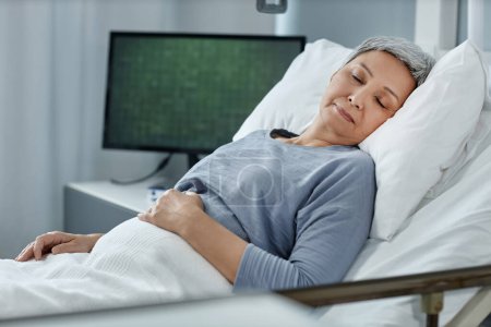 Foto de Mujer mayor que duerme en la cama en la sala de hospital durante su tratamiento con monitor que revisa los latidos de su corazón en segundo plano - Imagen libre de derechos