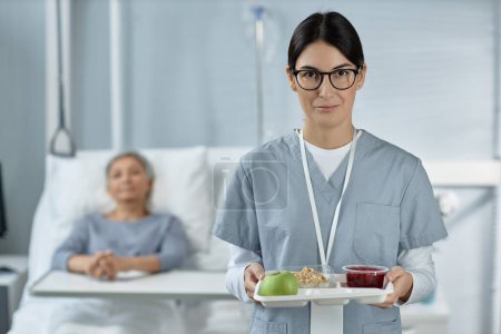Téléchargez les photos : Portrait d'une jeune infirmière en uniforme tenant un plateau avec de la nourriture et regardant la caméra, elle prend soin d'un patient malade dans un service hospitalier - en image libre de droit