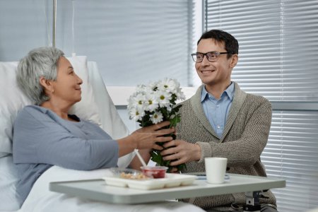 Téléchargez les photos : Fils rendant visite à sa mère aînée à l'hôpital pendant sa réhabilitation et lui donnant des fleurs - en image libre de droit