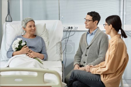 Téléchargez les photos : Femme malade âgée tenant un bouquet de fleurs et parlant à ses enfants adultes lors de leur visite à l'hôpital - en image libre de droit