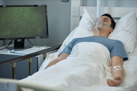 Téléchargez les photos : Jeune homme couché sur le lit avec masque à oxygène sur le visage pendant le traitement dans la salle d'hôpital - en image libre de droit