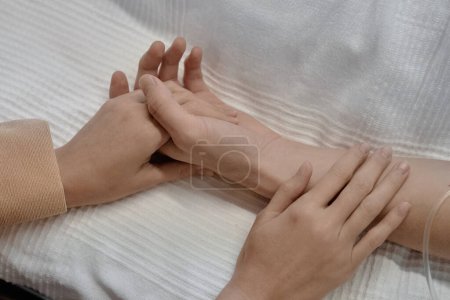 Téléchargez les photos : Gros plan de la jeune femme tenant la main de son mari alors qu'il était allongé sur le lit dans la salle d'hôpital - en image libre de droit
