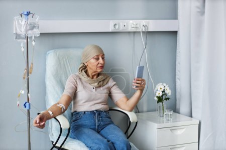 Foto de Retrato de una mujer mayor sentada en una silla cómoda con goteo intravenoso y usando un teléfono inteligente durante el tratamiento de quimioterapia - Imagen libre de derechos