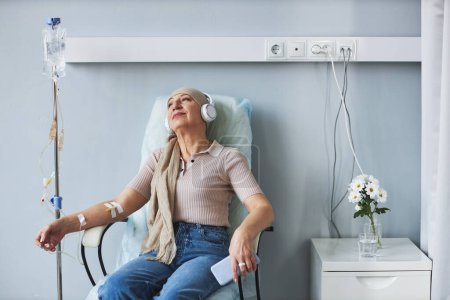 Foto de Retrato de una mujer mayor sentada en una silla cómoda con goteo intravenoso y escuchando música durante el tratamiento de quimioterapia, espacio para copiar - Imagen libre de derechos