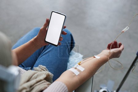 Photo for Close up of woman holding smartphone with white screen mockup during IV drip treatment, copy space - Royalty Free Image