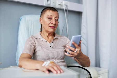Téléchargez les photos : Portrait de femme mature utilisant un smartphone et naviguant sur Internet pendant le traitement IV goutte à goutte, espace de copie - en image libre de droit
