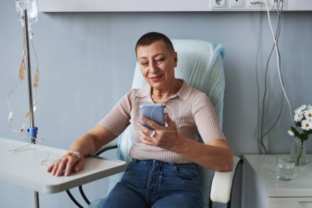 Foto de Retrato de una mujer madura sonriente usando un teléfono inteligente durante el tratamiento por goteo IV, espacio para copiar - Imagen libre de derechos