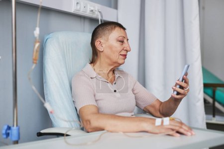 Foto de Retrato de vista lateral de una mujer madura sonriente usando un teléfono inteligente durante el tratamiento por goteo IV en la clínica, espacio para copiar - Imagen libre de derechos
