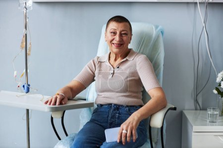 Foto de Retrato de una mujer madura sonriente mirando a la cámara durante el tratamiento por goteo intravenoso en la clínica, espacio para copiar - Imagen libre de derechos