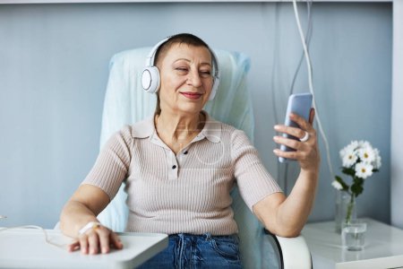 Foto de Retrato de una mujer mayor sonriente usando un teléfono inteligente con auriculares durante el tratamiento por goteo IV en el hospital - Imagen libre de derechos