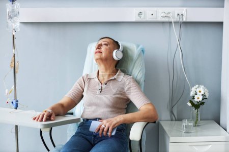 Téléchargez les photos : Portrait de femme âgée utilisant un casque relaxant pendant le traitement IV goutte à goutte à l'hôpital - en image libre de droit