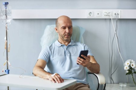 Foto de Retrato del hombre adulto usando teléfono inteligente durante el tratamiento por goteo IV en el hospital, espacio de copia - Imagen libre de derechos