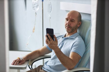 Foto de Retrato de hombre adulto calvo usando teléfono inteligente durante el tratamiento por goteo IV en la clínica, espacio de copia - Imagen libre de derechos