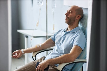 Photo for Side view portrait of adult man sitting in comfortable chair with IV drip during treatment session and relaxing - Royalty Free Image
