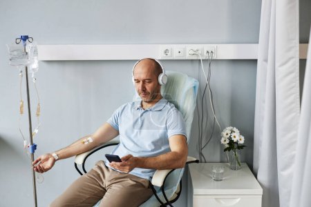 Foto de Retrato mínimo del hombre adulto usando smartphone con auriculares durante el tratamiento por goteo IV en la clínica - Imagen libre de derechos