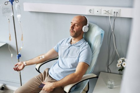 Foto de Retrato de alto ángulo del hombre adulto que usa auriculares y se relaja durante el tratamiento por goteo intravenoso en la clínica - Imagen libre de derechos