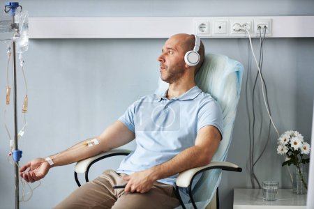 Foto de Retrato de vista lateral del hombre adulto que usa auriculares y escucha música durante el tratamiento por goteo intravenoso en la clínica - Imagen libre de derechos