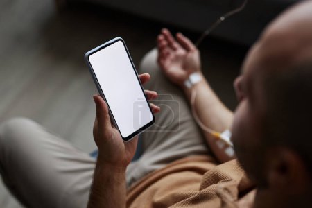 Foto de Primer plano del hombre adulto utilizando el teléfono inteligente con la maqueta de pantalla blanca durante el tratamiento por goteo IV - Imagen libre de derechos