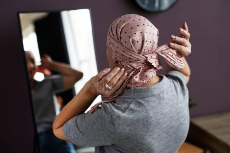 Foto de Vista posterior de una mujer calva irreconocible atando el pañuelo de la cabeza mirando en el espejo - Imagen libre de derechos