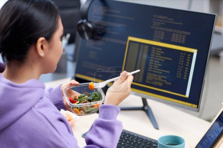 Téléchargez les photos : Gros plan du programmeur d'ordinateur féminin mangeant le déjeuner à emporter sur le lieu de travail tout en écrivant et en examinant le code - en image libre de droit