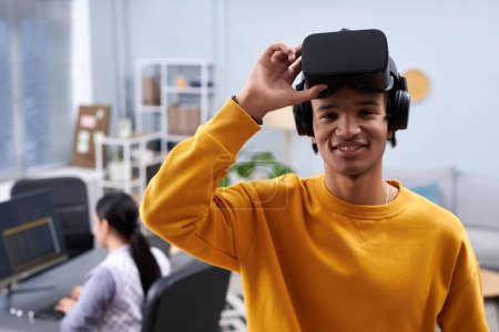 Waist up portrait of black young man using VR in IT development office and smiling at camera, copy space