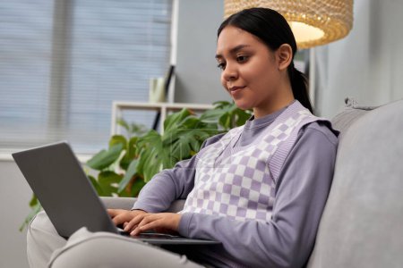 Téléchargez les photos : Portrait de jeune fille vue de côté à l'aide d'un ordinateur portable et souriant tout en se relaxant sur le canapé à la maison, espace de copie - en image libre de droit