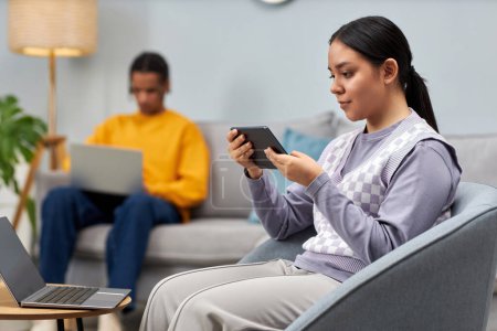 Téléchargez les photos : Portrait de jeune fille vue de côté à l'aide d'une tablette et d'un ordinateur portable assis sur le canapé dans le salon de bureau, espace de copie - en image libre de droit