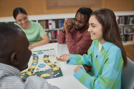 Foto de Retrato vista lateral de la joven sonriente jugando juego de mesa con un grupo de amigos en la biblioteca y divertirse juntos, espacio de copia - Imagen libre de derechos