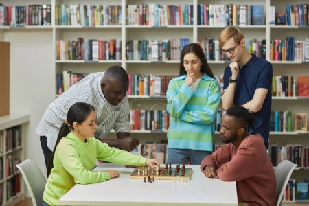 Téléchargez les photos : Vue latérale à deux jeunes gens jouant aux échecs dans la bibliothèque avec un groupe diversifié d'amis regardant - en image libre de droit