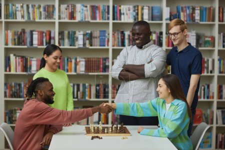 Téléchargez les photos : Vue latérale de deux joueurs d'échecs serrant la main sur la table avec un groupe diversifié de personnes regardant le match - en image libre de droit