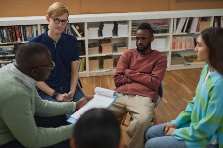 Foto de Vista de ángulo alto en un grupo diverso de jóvenes sentados en círculo durante la sesión de terapia en la universidad con el psicólogo masculino - Imagen libre de derechos