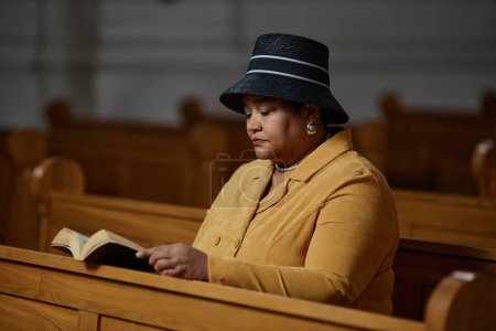 Elegante mujer madura en sombrero visitando la vieja iglesia, ella sentada en el banco y leyendo la oración en la Biblia