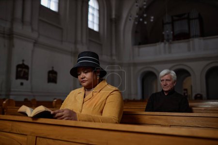 Elegant woman in hat reading Bible sitting on wooden bench in church with senior man sitting behind her