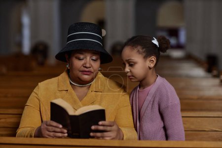 Téléchargez les photos : Grand-mère lisant la Bible à sa petite-fille alors qu'ils étaient assis sur le banc à l'église - en image libre de droit
