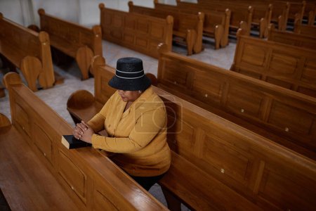 Foto de Vista de ángulo alto de la elegante mujer madura sentada en el banco con la Biblia y orando durante el ministerio en la iglesia - Imagen libre de derechos