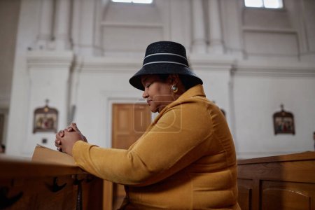 Téléchargez les photos : Vue latérale de la femme dodue afro-américaine assise sur un banc avec la Bible et priant à l'église - en image libre de droit