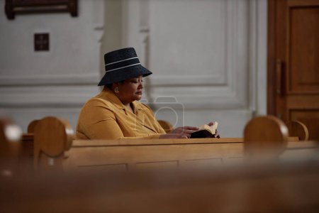 Foto de Mujer afroamericana en ropa elegante leyendo la Biblia mientras está sentada en el banco en la iglesia - Imagen libre de derechos