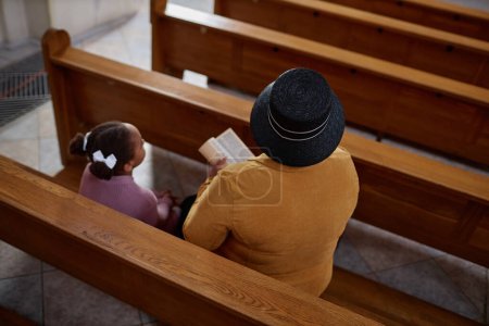 High angle view of grandmother reading Bible while sitting on bench with her granddaughter in church, they praying together