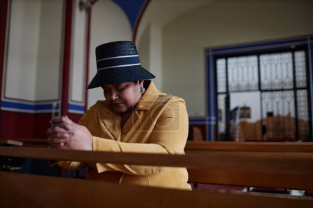 Téléchargez les photos : Femme élégante en chapeau assise sur un banc les yeux fermés et priant à l'église - en image libre de droit