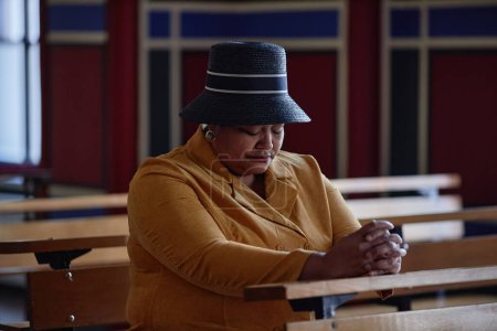 Téléchargez les photos : Femme afro-américaine au chapeau élégant priant les yeux fermés assis sur un banc à l'église - en image libre de droit