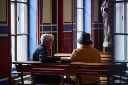 Téléchargez les photos : Vue arrière du prêtre aîné parlant à un visiteur assis sur un banc pendant son ministère à l'église - en image libre de droit