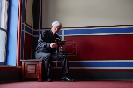 Téléchargez les photos : Prêtre principal lisant la Bible tout en se reposant sur le banc près de la fenêtre dans la vieille église - en image libre de droit