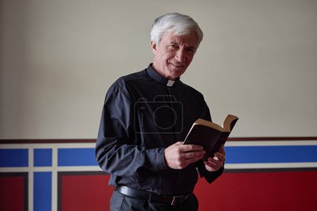 Foto de Retrato del sacerdote mayor sonriendo a la cámara mientras lee la oración en el libro de la Biblia - Imagen libre de derechos