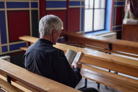 Foto de Vista trasera del sacerdote mayor leyendo la Biblia durante la oración mientras está sentado en el banco de la iglesia - Imagen libre de derechos