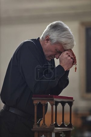 Téléchargez les photos : Image verticale d'un homme âgé avec des perles rôties priant debout derrière l'autel dans une église - en image libre de droit