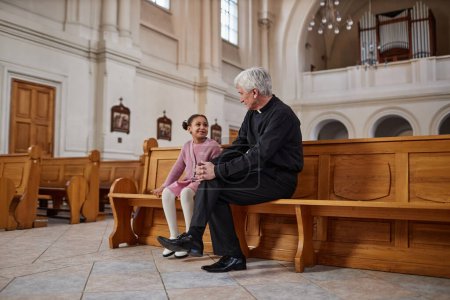 Photo for Senior priest talking to African American little girl while they sitting on bench in old church - Royalty Free Image