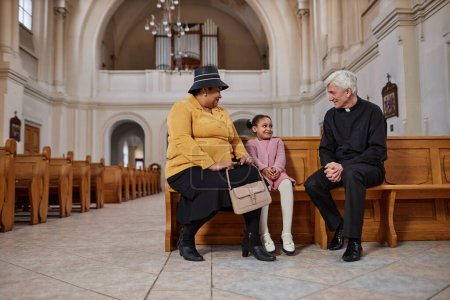 Téléchargez les photos : Afro-américaine grand-mère parler avec prêtre avec sa petite-fille alors qu'ils sont assis sur le banc dans l'église - en image libre de droit