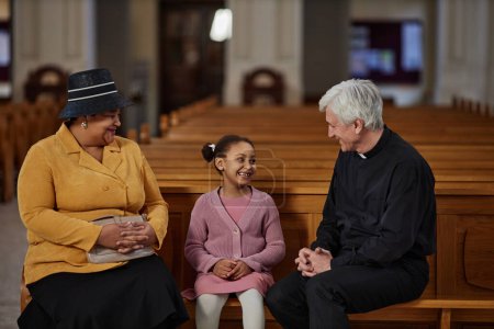 Téléchargez les photos : Petite fille parlant au prêtre alors qu'ils étaient assis sur le banc lors de sa visite à l'église avec sa grand-mère - en image libre de droit